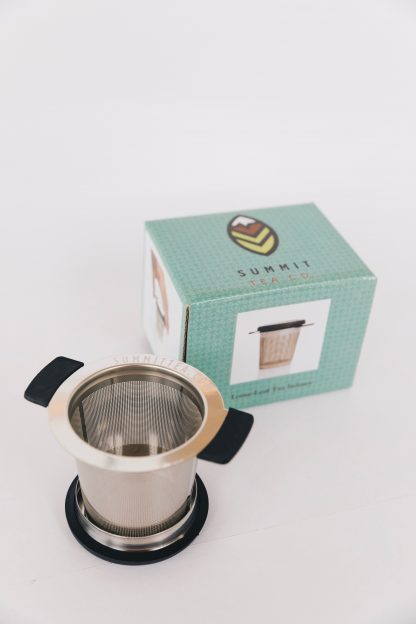 Stainless steel loose leaf tea infuser with black silicon support wings and lid/saucer with teal gift box on white background